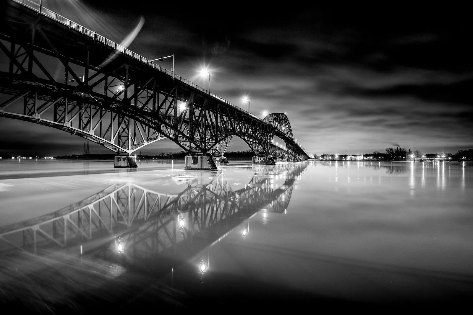Black-and-white photograph of a steel bridge and its reflection in still water beneath a dramatic night sky.