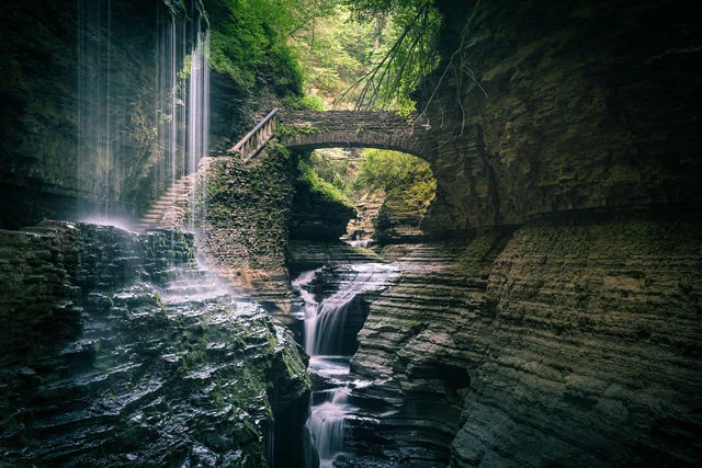 A waterfall flowing through a green, layered rock gorge beneath an arched stone bridge.