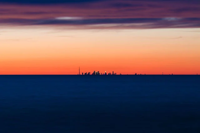 A vivid orange sunset above deep blue water, with a distant skyline along the horizon.
